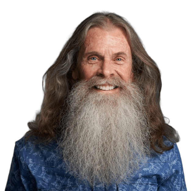 A smiling man with long, wavy gray hair and a full, bushy beard is wearing a blue shirt. The background is plain, emphasizing the subject's cheerful expression.