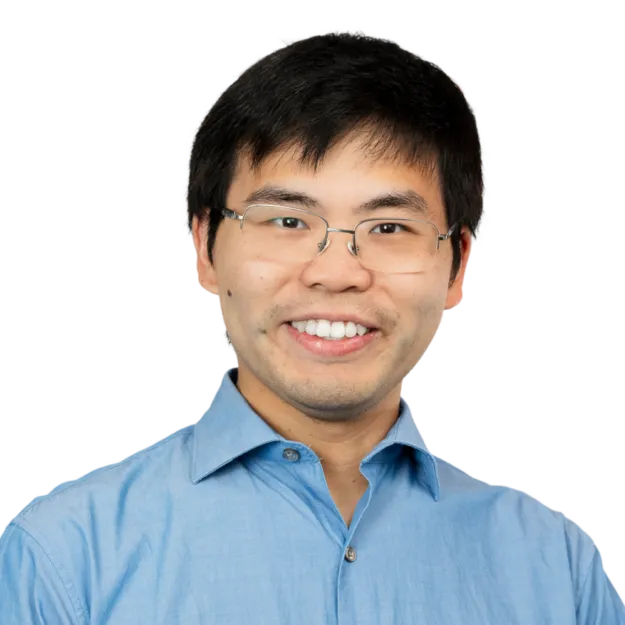 A person with short black hair, glasses, and a light blue button-up shirt is smiling at the camera against a plain white background.