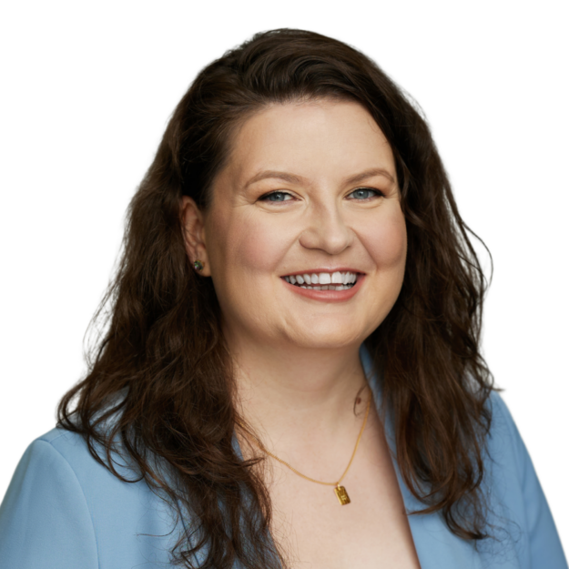 A woman with long, wavy brown hair is smiling. She is wearing a blue blazer, a light-colored top, and a gold necklace with a rectangular pendant. The background is plain white.
