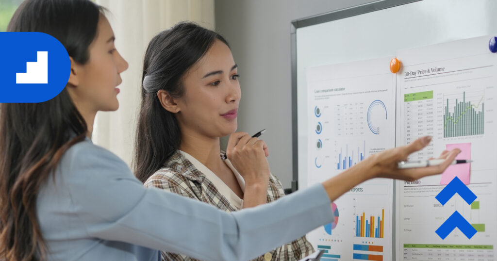 Professionals in a modern office having a discussion around a table, with charts and data displayed on a large screen.