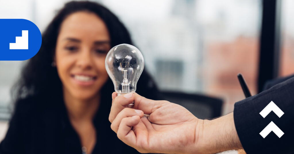 Close-up of a person holding a clear light bulb in focus while a smiling woman in the background looks on, symbolizing new ideas and innovation.