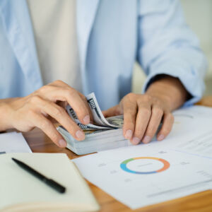 Person counting a stack of hundred-dollar bills at a desk with financial documents, charts, and a pen nearby.