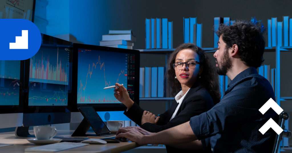 A woman and a man sit together in front of large monitors displaying data visualizations and charts, discussing insights in a modern office filled with bookshelves.