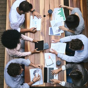 High-angle view of a diverse group of business professionals seated around a wooden table, collaborating with charts, printed reports, a laptop, a tablet, a calculator, and coffee cups during a meeting.