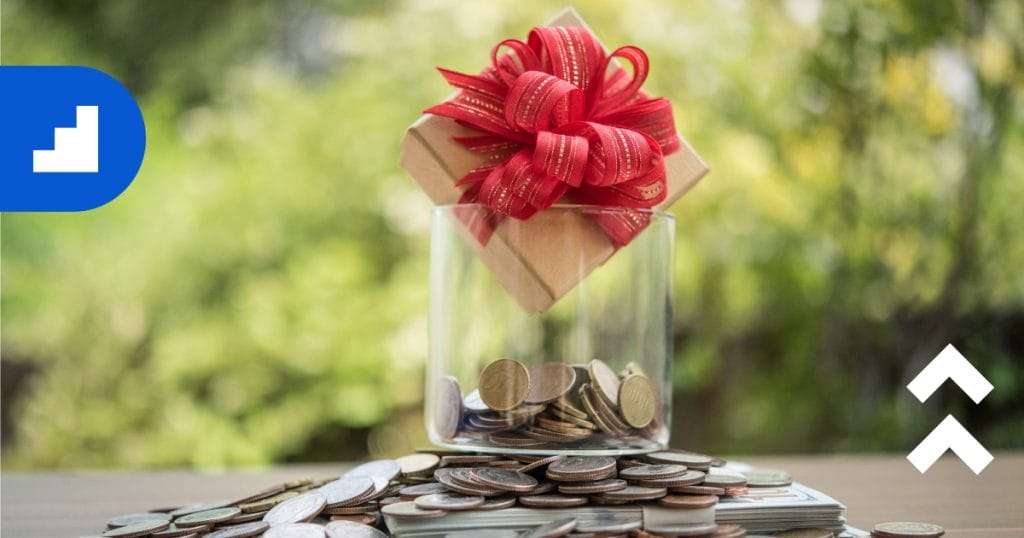 A glass jar filled with stacked coins sits on a table outdoors, with a wrapped gift box tied with a red ribbon placed on top, symbolizing donations, fundraising, and generosity.
