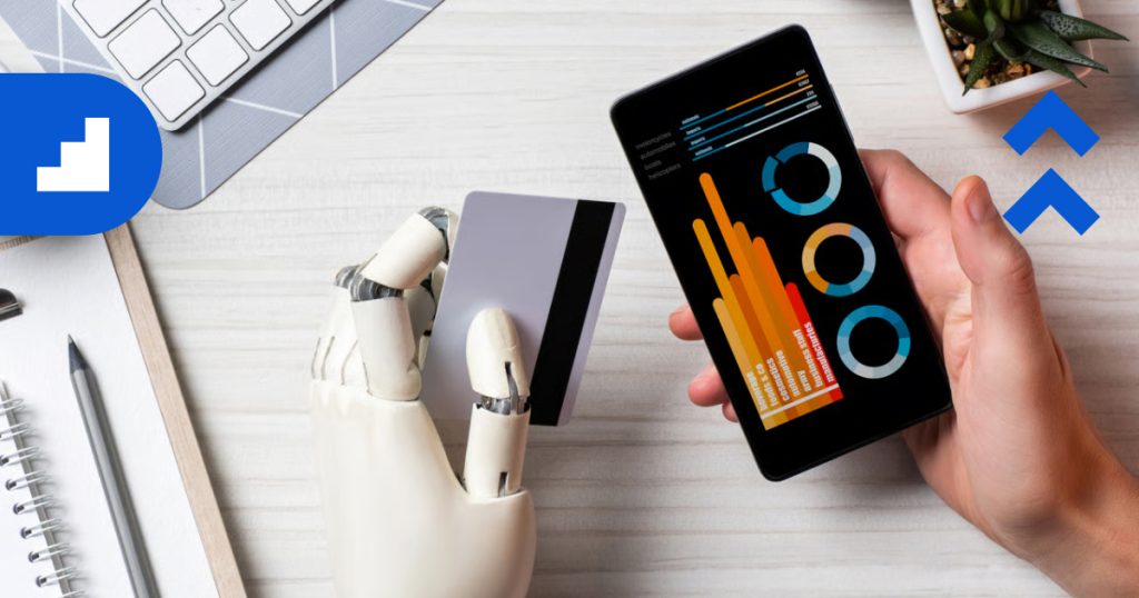 The background shows a desk with a laptop, a smartphone displaying colorful data charts, and a robotic hand holding a credit card. In the top corners, there are blue graphic elements: a stepped icon on the left and two upward-pointing chevrons on the right.