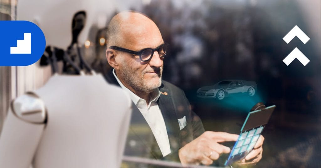 An older man with glasses and a beard looks thoughtfully at a tablet device. Overlaid on the image is a holographic projection of a sports car. The image also features a blue logo in the top left and a white double-arrow icon in the top right.