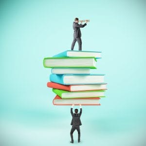 A conceptual illustration against a teal background showing one businessman at the bottom holding up a towering stack of colorful books in green, red, and blue, while another businessman in a suit stands on top looking through a brass telescope, symbolizing teamwork, knowledge, vision, and leadership.