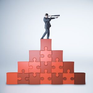 A businessman stands atop a pyramid of red puzzle pieces, looking through a telescope and pointing forward against a plain grey background.