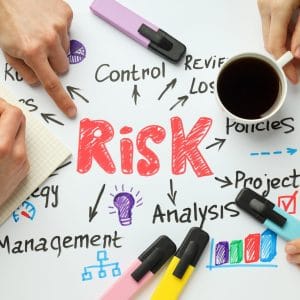 Multiple hands collaborating around a white surface with "Risk" written in large red letters at the center, surrounded by hand-drawn icons and related keywords including "Management," "Control," "Analysis," "Policies," "Project," and "Strategy," with colorful markers, a notepad, and a cup of coffee, illustrating a brainstorming session on risk management concepts.