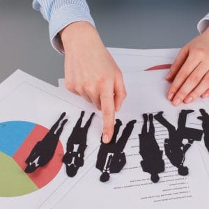 A person in a blue striped shirt pointing to one of several black paper cutout silhouettes of business figures laid out on a desk covered with documents, charts, and a pie chart, representing the concept of employee selection or recruitment.