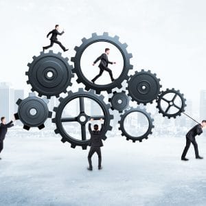 A conceptual business illustration showing five suited businessmen interacting with a large interconnected system of dark metal gears against a city skyline backdrop, with one pushing a gear from the left, one holding up the central gear, one pulling a rope on the right, and two others running on top of and inside the gears, symbolizing teamwork, collaboration, and the mechanics of business operations.