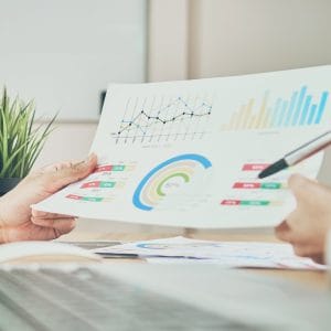 Two people in a business meeting reviewing printed data reports featuring line graphs, bar charts, and circular infographics, with one person holding up the document and another pointing to it with a pen, at a wooden desk with a laptop and small potted plant in the background.