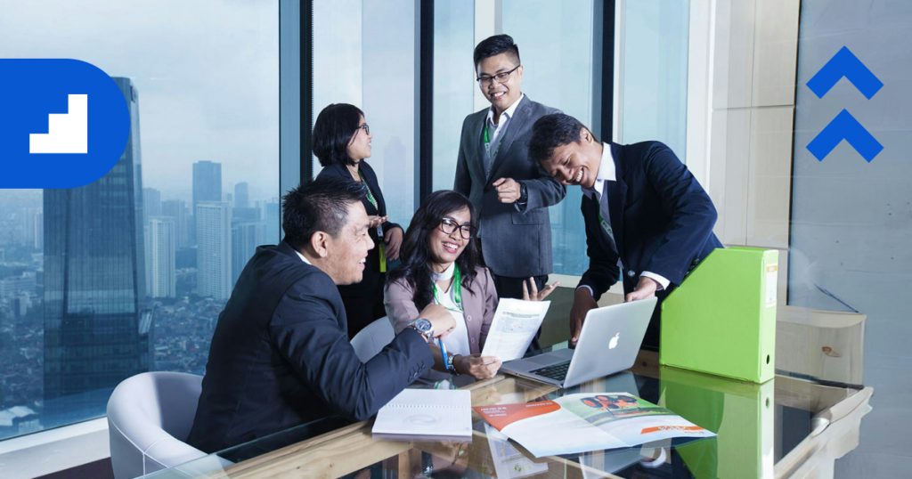 Four professionally dressed colleagues are gathered around a table in a modern office, smiling and discussing paperwork.