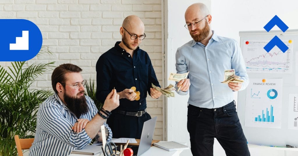 Two men in an office count revenue beside a whiteboard with charts and graphs. One holds dollar bills and a notebook, while the other has coins.
