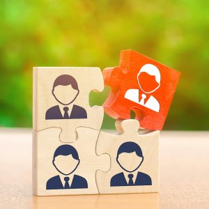 Four wooden puzzle pieces with suit-and-tie icons are being assembled against a blurred green background. Three tan pieces are connected at the base, while a fourth bright orange piece is being placed on top, representing a strategic hire or the final piece of a team.