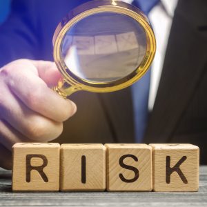Close-up of a person in a business suit holding a magnifying glass over wooden blocks that spell the word 'RISK.' The magnifying glass focuses on the letters, symbolizing risk assessment, due diligence, and careful analysis.