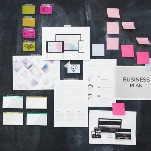 A top-down view of a dark brainstorm board featuring a "Business Plan" document, digital UI mockups, marketing strategies, and various colorful sticky notes.