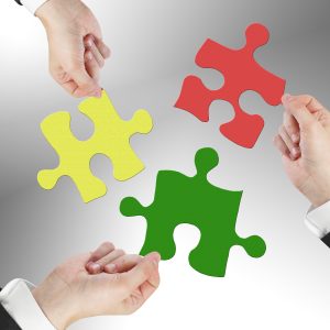 Four hands in business attire hold large, colorful puzzle pieces (yellow, red, and green) against a grey gradient background, symbolizing teamwork and collaboration.