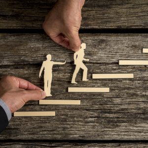 A top-down view of hands placing paper cutouts of people on a wooden staircase. One hand assists a figure climbing the steps while another holds a figure at a lower level, symbolizing mentorship, career progression, and leadership support.