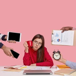 An overwhelmed woman sits at a desk while multiple hands thrust a tablet, phone, clipboard, and alarm clock toward her, symbolizing burnout and excessive workload.