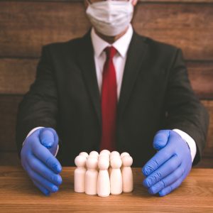 A person in a business suit, wearing a protective face mask and blue gloves, cups their hands around a group of small wooden figures on a wooden table, symbolizing employee protection, workplace safety, and risk management.