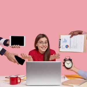 A woman in a red polka-dot shirt sits at a laptop, shrugging and smiling as hands offer work tools—perfectly illustrating the need for a Workplace Strategy Call (60 minutes)—with a pink background.