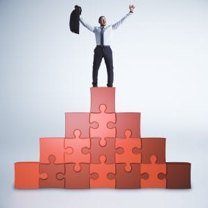 A celebratory businessman in a white shirt and tie stands triumphantly at the top of a pyramid made of large, red and orange puzzle pieces. He has his arms raised in the air, holding his suit jacket in one hand, signifying success and the completion of a complex goal.