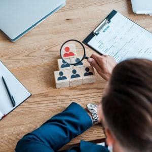 Top-down view of a professional in a suit using a magnifying glass to examine a pyramid of wooden blocks. One block is highlighted in red, representing a specific candidate, alongside a resume, laptop, and notebook on a wooden desk, symbolizing recruitment and talent selection.