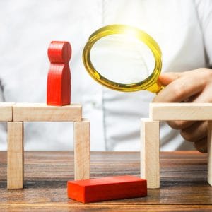 A person holding a gold magnifying glass over a gap between wooden blocks structured like a bridge. A red wooden figure stands on one side of the gap, while a fallen red block lies below, symbolizing strategic analysis, gap assessment, and identifying organizational risks or bottlenecks.