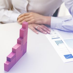 A close-up view of a businesswoman in a white dress shirt sitting at a desk with her hands crossed, looking at a rising staircase chart built from pink wooden blocks. An analytical financial report rests on the table next to the blocks, illustrating data evaluation, step-by-step scaling, and building a foundation for sustainable expansion during a Growth Strategy Consulting session.