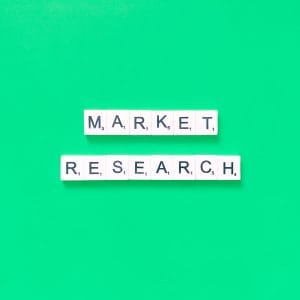 A top-down, minimalist view of white letter tiles spelling out 'MARKET RESEARCH' arranged in two parallel rows centered on a solid vibrant green background. The clean and organized layout symbolizes target audience analysis, systematic insights, and data-driven foundational planning necessary for building a Brand Positioning & Go-To-Market Strategy.