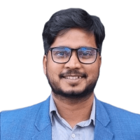 A person with short dark hair, a beard, and glasses is smiling at the camera. They are wearing a blue blazer over a light-colored button-down shirt. The background is transparent.